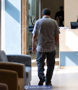 بیمار میانسال در حال ورود به کلینیک قلب با فضای انتظار راحت، صندلی‌های نرم، میز پذیرش در محیطی روشن با رنگ‌های آبی و سفید.